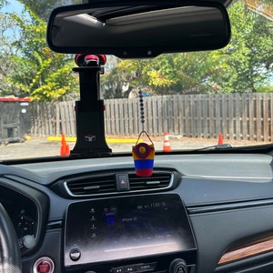 May include: A black car interior with a black dashboard and a screen displaying "iPhone (2)" and "Track". There is a red and black phone holder attached to the rearview mirror and a small colorful woven hanging ornament.