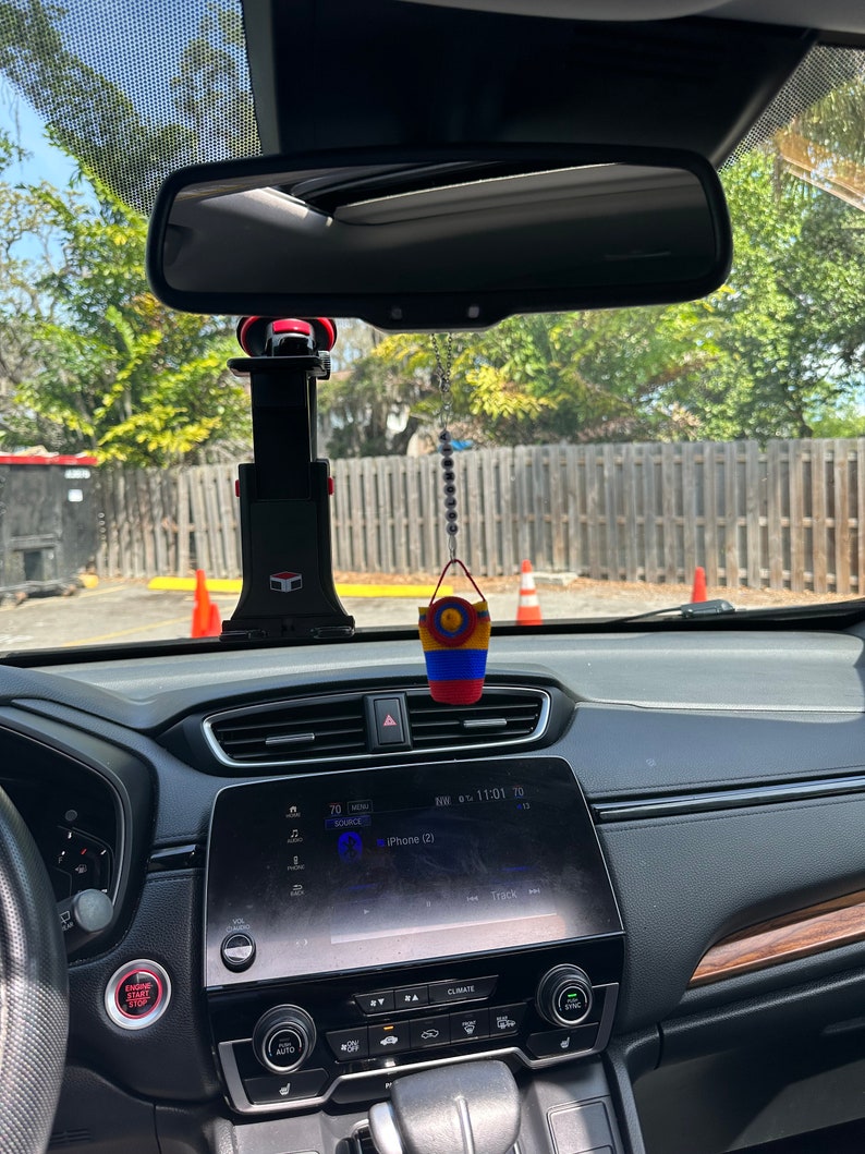 May include: A black car interior with a black dashboard and a screen displaying "iPhone (2)" and "Track". There is a red and black phone holder attached to the rearview mirror and a small colorful woven hanging ornament.