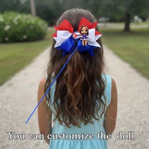 May include: A hair bow featuring layered red, white, and blue ribbons with a circular Cuban flag emblem and a small doll figurine attached. The text at the bottom of the image states: You can customize the doll.