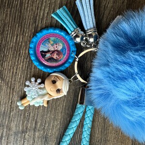 May include: A blue fuzzy keychain with a silver key ring, a blue and pink character charm, and a blue and white character charm. The keychain also has two blue tassels.