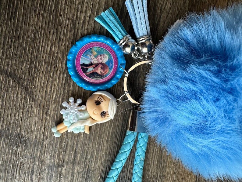 May include: A blue fuzzy keychain with a silver key ring, a blue and pink character charm, and a blue and white character charm. The keychain also has two blue tassels.