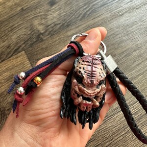 May include: A black leather keychain with a Predator character charm. The charm is made of a brown and beige material and has a detailed face with dreadlocks. The keychain also has a red, black, and blue braided cord with silver and gold beads.