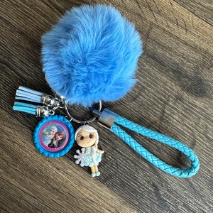 May include: A blue fuzzy keychain with a small blue and pink charm featuring a picture of two girls. The charm is attached to a blue braided strap.
