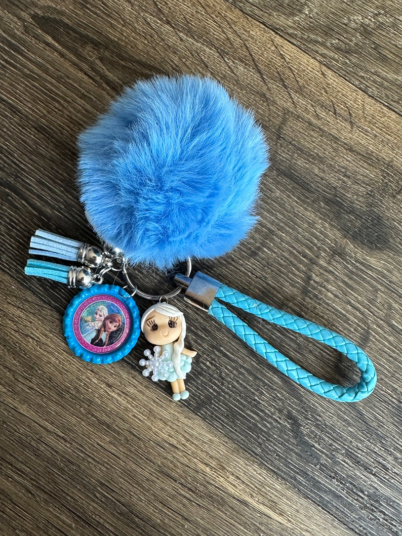 May include: A blue fuzzy keychain with a small blue and pink charm featuring a picture of two girls. The charm is attached to a blue braided strap.