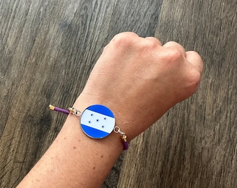 May include: A purple cord bracelet with a round charm featuring the flag of Honduras. The charm is silver with a blue background and five white stars.
