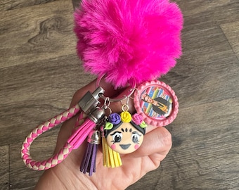 May include: A pink fuzzy keychain with a braided leather strap, a pink and white tassel, a purple tassel, a yellow tassel, a round pink charm with a picture of a man, and a round charm with a cartoon image of a woman with a flower crown.