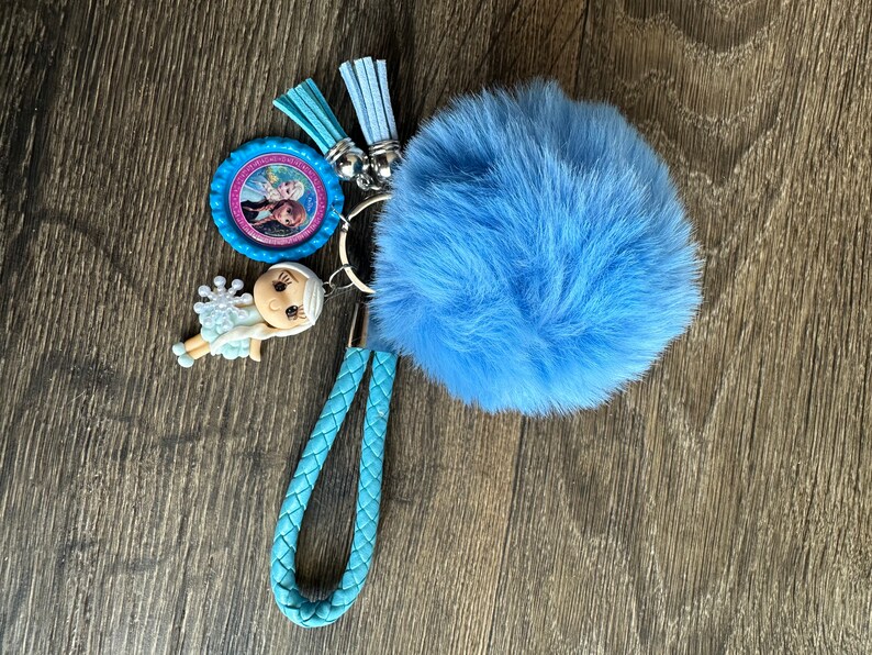 May include: A blue fuzzy keychain with a braided blue strap, a small plastic doll, and a round charm with a picture of two girls.