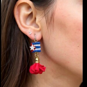 May include: A close-up of a dangling earring featuring a Cuban flag design. The earring has a blue and white flag with a red star, a gold-colored chain, and a red tassel. The earring is attached to a silver hoop.