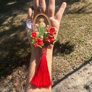 May include: A red tassel hangs from a gold oval frame with a religious image inside. The frame is surrounded by red roses.