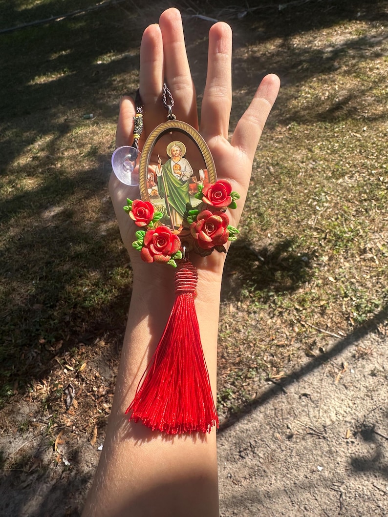 May include: A red tassel hangs from a gold oval frame with a religious image inside. The frame is surrounded by red roses.