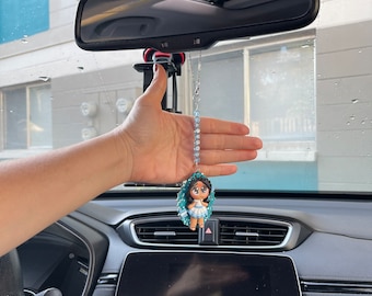 May include: A blue and white car charm featuring a girl with a crown and a blue dress. The charm is hanging from a rearview mirror in a car.