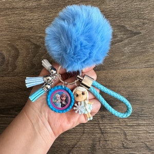 May include: A blue fuzzy keychain with a blue and pink character charm, a blue braided strap, and a silver ring. The charm features a picture of two girls with blonde hair.