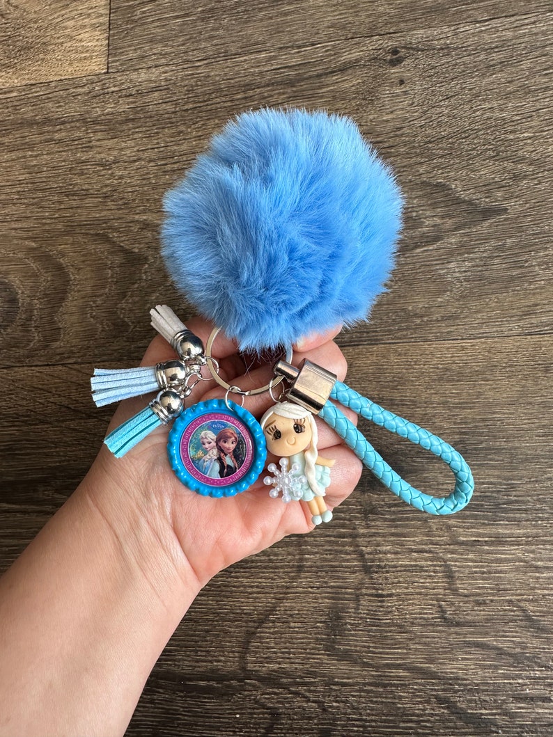 May include: A blue fuzzy keychain with a blue and pink character charm, a blue braided strap, and a silver ring. The charm features a picture of two girls with blonde hair.