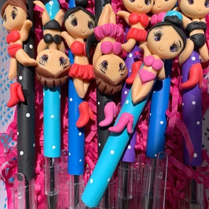 May include: A collection of pens with colorful barrels, each adorned with a miniature figurine of a woman in a bikini. The pens have white polka dots and are in various colors, including black, blue, and purple. The figurines are in different poses.