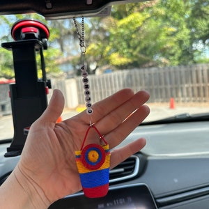 May include: A rearview mirror charm featuring a small crocheted bag in the colors of the Colombian flag, yellow, blue, and red, with a yellow and red circle on the top. The bag is attached to a silver chain with white beads spelling out "COLOMBIA".