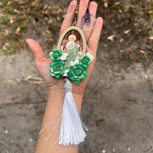 May include: A green and gold car charm featuring a religious figure with a white tassel. The charm is surrounded by green and white flowers.