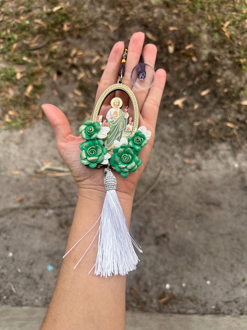 May include: A green and gold car charm featuring a religious figure with a white tassel. The charm is surrounded by green and white flowers.