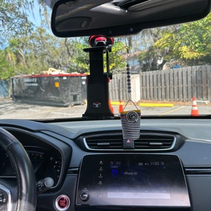 May include: A black car interior with a red button labeled "Engine Start Stop" and a black and white striped car air freshener hanging from the rearview mirror. The car's infotainment system displays "iPhone (2)" and the time is 11:03.