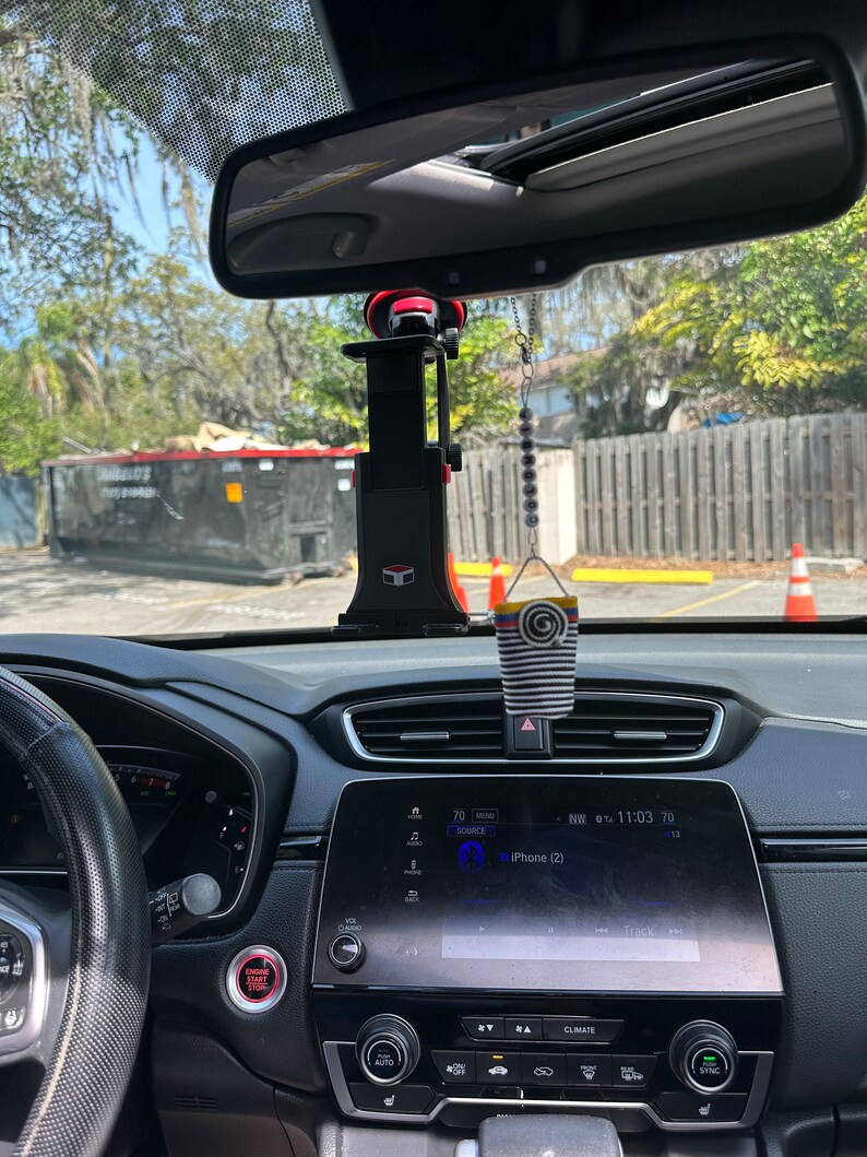 May include: A black car interior with a red button labeled "Engine Start Stop" and a black and white striped car air freshener hanging from the rearview mirror. The car's infotainment system displays "iPhone (2)" and the time is 11:03.