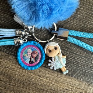 May include: A blue fuzzy keychain with a blue and pink character charm featuring Elsa and Anna from Frozen. The charm is attached to a silver key ring and a blue braided cord.
