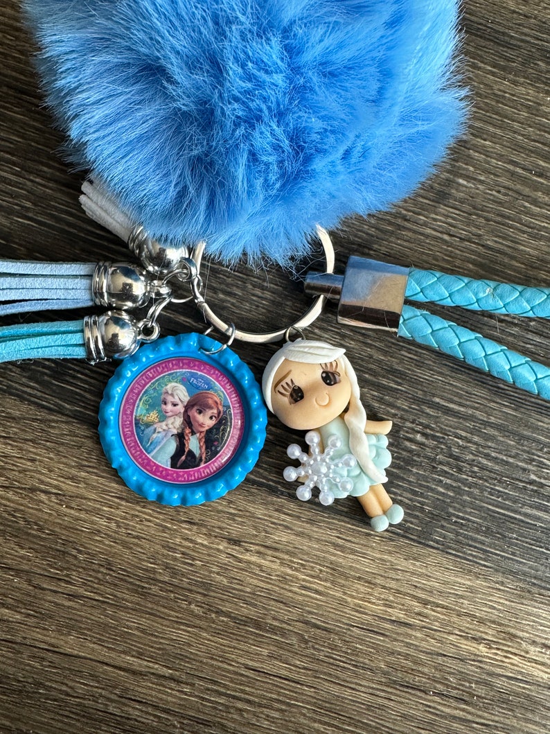 May include: A blue fuzzy keychain with a blue and pink character charm featuring Elsa and Anna from Frozen. The charm is attached to a silver key ring and a blue braided cord.