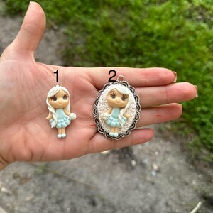 May include: Two handmade polymer clay charms featuring a girl with blonde hair and a light blue dress. The charm on the left is a simple charm, while the charm on the right is set in a silver oval frame with a decorative edge.