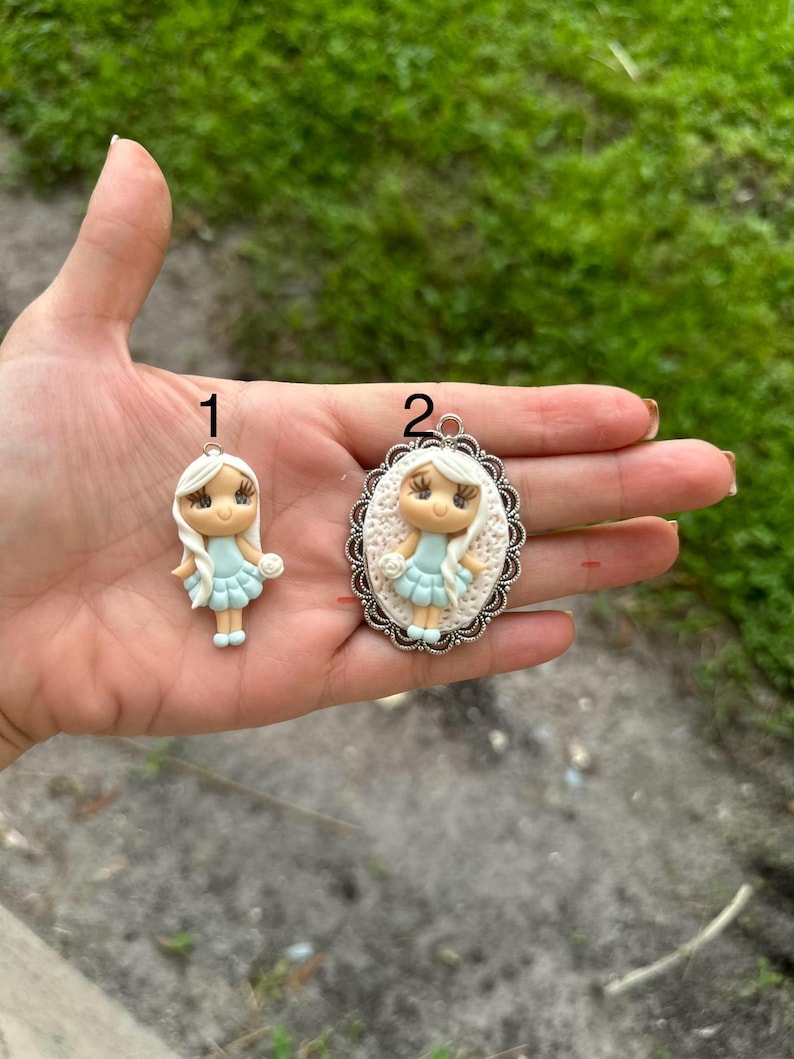 May include: Two handmade polymer clay charms featuring a girl with blonde hair and a light blue dress. The charm on the left is a simple charm, while the charm on the right is set in a silver oval frame with a decorative edge.