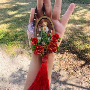 May include: Oval-shaped car charm with a picture of a saint, surrounded by red roses and a red tassel.