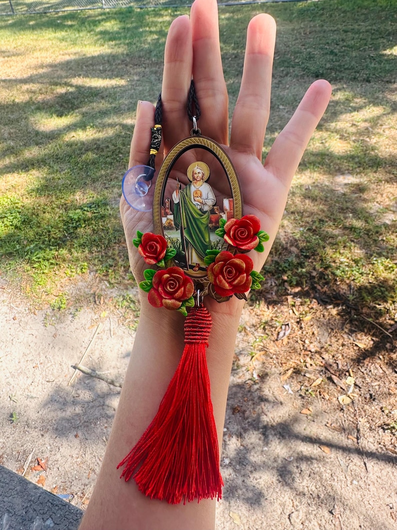 May include: Oval-shaped car charm with a picture of a saint, surrounded by red roses and a red tassel.