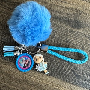 May include: A blue fuzzy keychain with a blue braided strap and a round charm featuring a picture of two girls from the movie Frozen. The charm is attached to a silver ring with a small blue tassel.