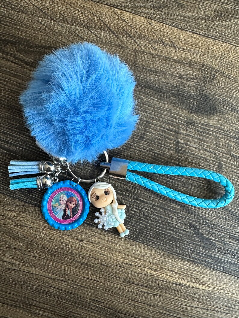 May include: A blue fuzzy keychain with a blue braided strap and a round charm featuring a picture of two girls from the movie Frozen. The charm is attached to a silver ring with a small blue tassel.
