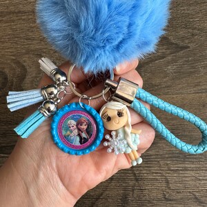 May include: A blue fuzzy keychain with a silver key ring, two blue tassels, and a blue braided strap. A small blue and pink plastic charm with a picture of Elsa and Anna from Frozen is attached to the key ring. A small doll with a white dress and a snowflake in her hand is attached to the braided strap.