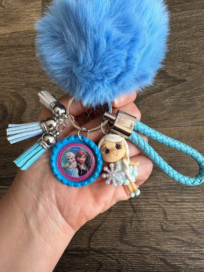 May include: A blue fuzzy keychain with a silver key ring, two blue tassels, and a blue braided strap. A small blue and pink plastic charm with a picture of Elsa and Anna from Frozen is attached to the key ring. A small doll with a white dress and a snowflake in her hand is attached to the braided strap.