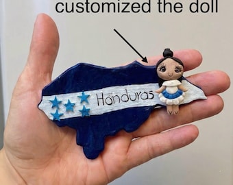 May include: A handcrafted Honduras-shaped ornament in blue and white, featuring five blue stars and the word "Honduras." A small doll in a white and blue dress is attached. The text "You can customized the doll" is also present.