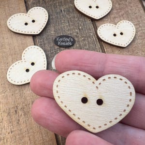 May include: A set of four unfinished wooden heart-shaped buttons with two holes each. The buttons have a stitched edge detail.