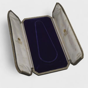 Op de afbeelding: Een open, vintage sieradendoosje met een donkerblauwe fluwelen binnenkant en een goudkleurige rand. De doos is gevoerd met crèmekleurig satijn en heeft een scharnierend ontwerp. De buitenkant is donker met gouden accenten.