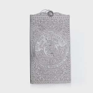 May include: A silver-colored, rectangular calendar with ornate engraved designs. The calendar features a central circular dial with text and numbers, surrounded by scrolling floral patterns. The top has a small loop for hanging.