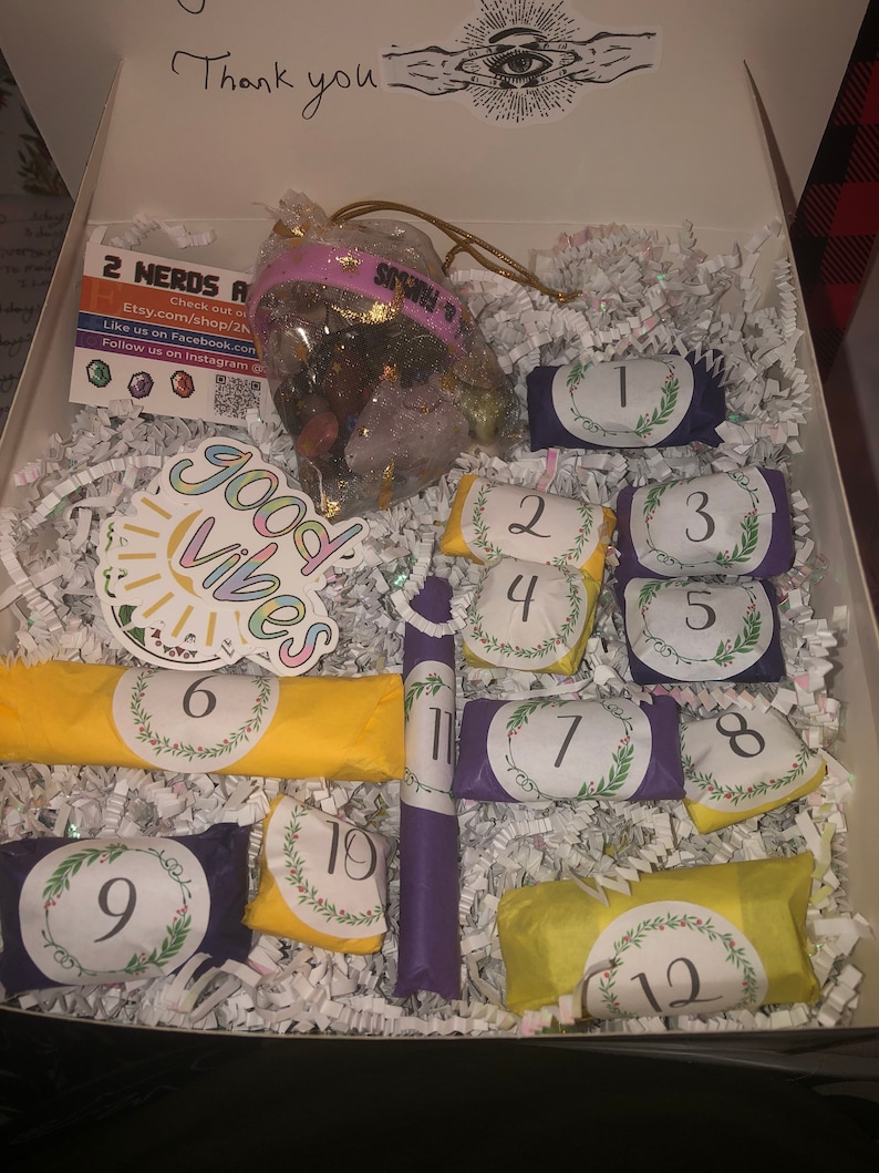 May include: An open white box filled with shredded paper, containing a bag of crystals, numbered wrapped items, and a sticker that says "Good Vibes". The box also has the text "Thank you".