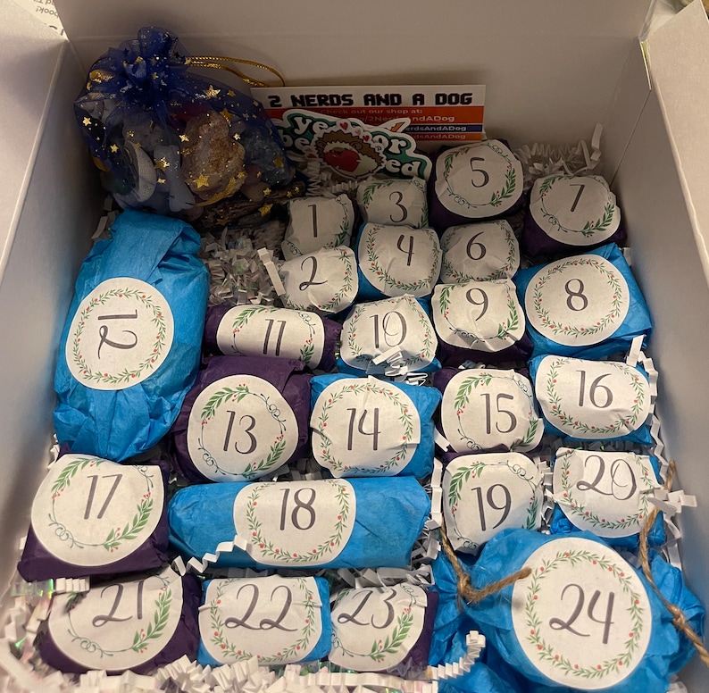 May include: An open box filled with wrapped gifts for an advent calendar. Each gift is wrapped in blue or purple paper and labeled with a number from 1 to 24. A small bag with a gold drawstring is visible in the upper left corner. The text "2 Nerds and a Dog" is visible.