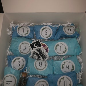 May include: An open white box filled with light blue wrapped packages, each labeled with a number from 1 to 12. The packages are decorated with a wreath design. A small glass bottle with stones and stickers are also visible.