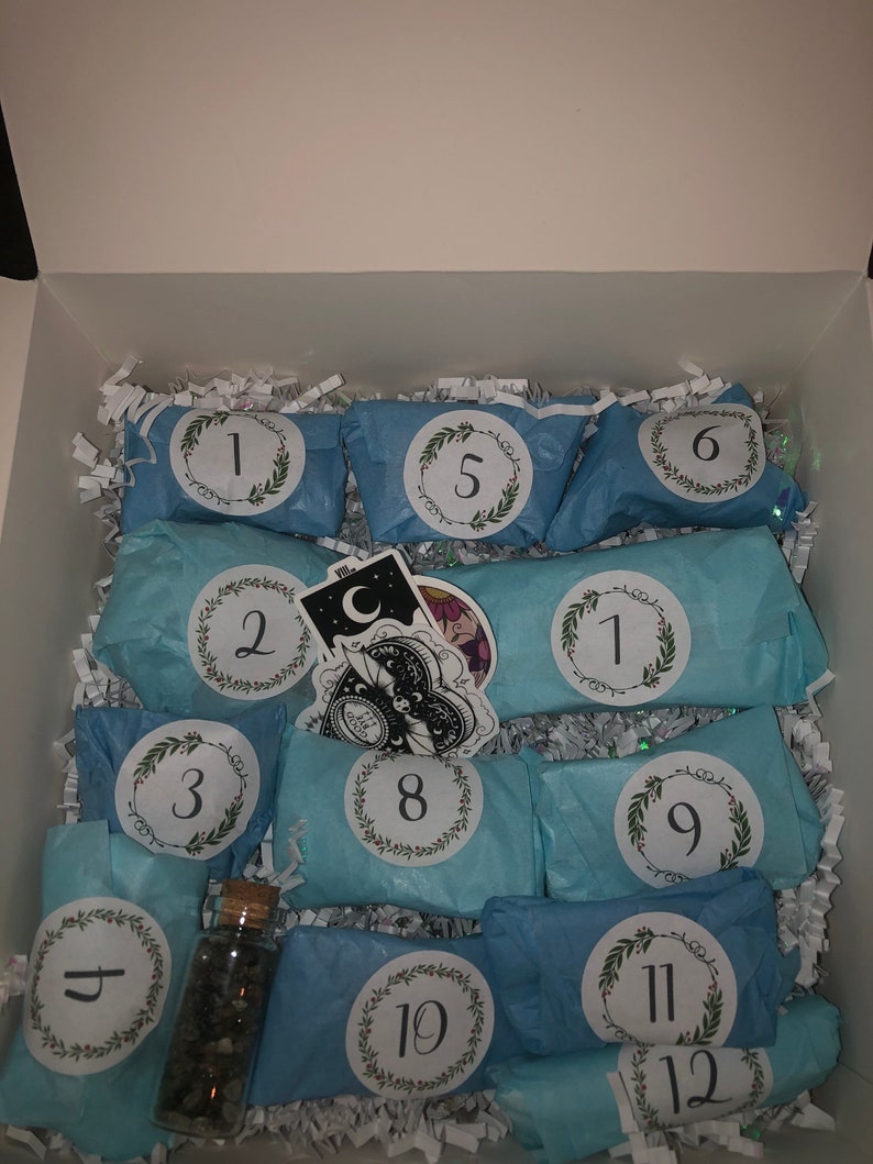 May include: An open white box filled with light blue wrapped packages, each labeled with a number from 1 to 12. The packages are decorated with a wreath design. A small glass bottle with stones and stickers are also visible.