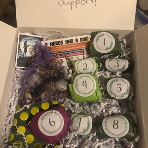 May include: An open white box filled with gifts, including eight numbered packages wrapped in green and purple paper. The box contains a variety of items, including a card with the text "2 Nerds and a Dog" and the words "Thank you for your Support!"
