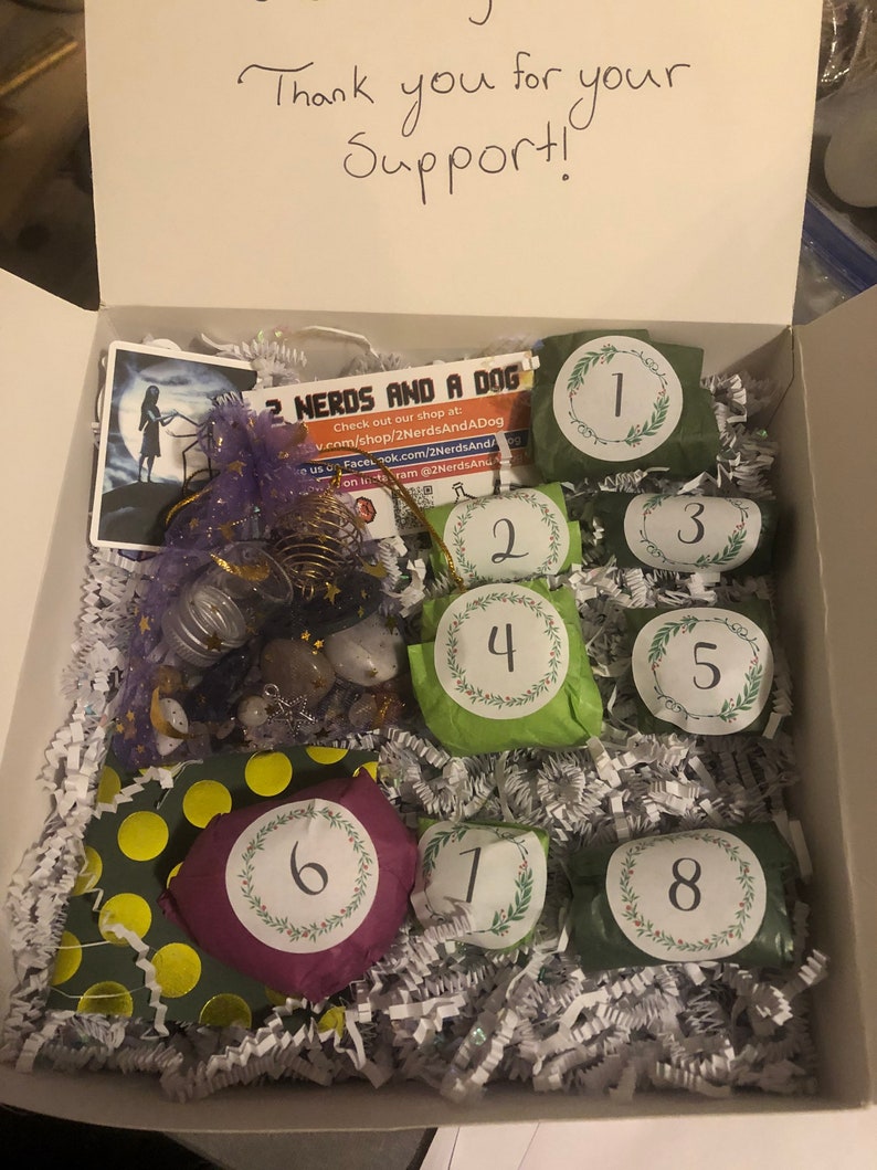 May include: An open white box filled with gifts, including eight numbered packages wrapped in green and purple paper. The box contains a variety of items, including a card with the text "2 Nerds and a Dog" and the words "Thank you for your Support!"