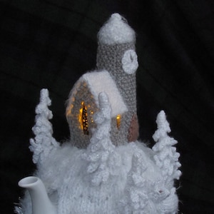 Könnte beinhalten: Eine weiße Keramik-Teekanne, verziert mit einer gestrickten Winterszene. Das gemütliche Design zeigt eine Kirche mit Glockenturm, schneebedeckte Bäume und eine flauschige, weiße Strickbasis. Silberner Lametta sorgt für einen festlichen Touch.