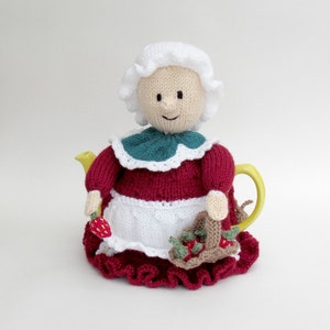 Op de afbeelding: Een gebreide theepotwarmer in de vorm van een vrouw die een rode jurk, een witte schort en een witte muts draagt. Ze houdt een mandje met aardbeien vast.