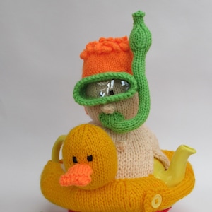 May include: A knitted teapot cozy shaped like a yellow duck with an orange beak and green flippers. A knitted person wearing an orange hat, green goggles, and a green snorkel sits on top of the duck.