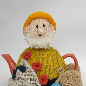 Może przedstawiać: Wełniany pokrowiec na czajnik w kształcie osoby w żółtym kapeluszu i białym brodzie. Pokrowiec ma czerwone i pomarańczowe akcenty oraz przymocowaną z boku dzianą torbę.