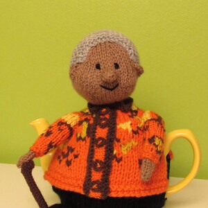 Pode incluir: Uma capa de chá de tricô em forma de pessoa vestindo um suéter marrom e laranja com uma echarpe marrom e calças pretas. A pessoa está segurando uma bengala.