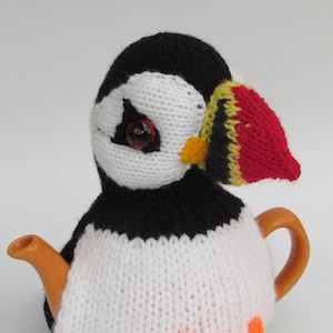 Könnte beinhalten: Ein gestrickter Teekannenwärmer in Form eines Papageientauchers mit orangenen Füßen, einem schwarz-weißen Körper und einem rot-gelb-schwarzen Schnabel.