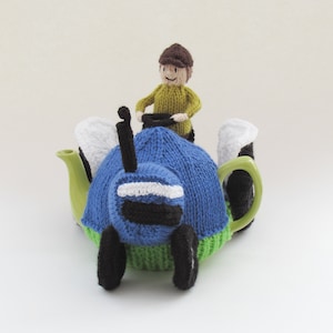 May include: A knitted blue and green tractor teapot cozy with a knitted person figure driving the tractor. The tractor has black wheels and a white stripe on the front.
